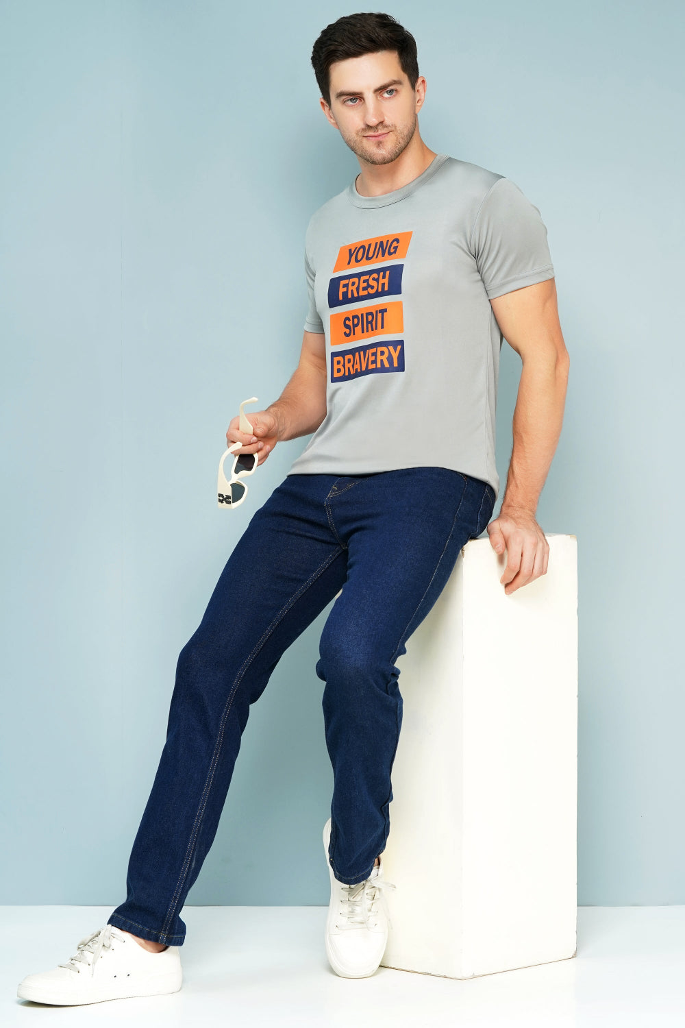 Man wearing a gray t-shirt with colorful text and blue jeans, sitting on a white block against a light blue background.