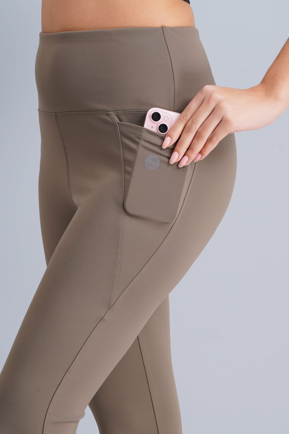 Person wearing brown leggings with a pocket holding a phone on a plain background